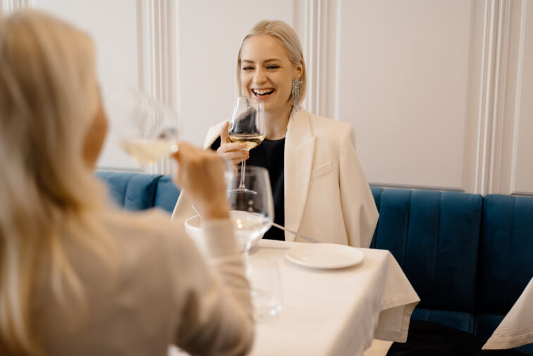 Cheerful blond woman in elegant clothes raising glass of white wine and smiling while having lunch with girlfriend in hotel restaurant.