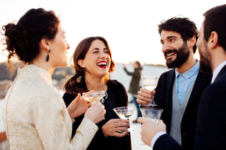 Anonymous cheerful colleagues with glasses of alcoholic drinks speaking during festive event on rooftop and looking at each other