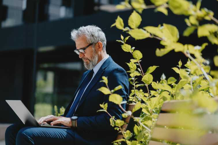 Senior businessman in a suit working on a laptop outdoors in an urban garden, surrounded by greenery on a sunny day.